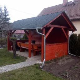 Drewniana altana z ciemnym dachem i rynnami, wyposażona w stół i ławy, usytuowana w ogrodzie z kostką brukową i zielonym trawnikiem.