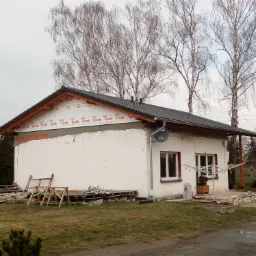 Dom w trakcie prac: widoczna nowa, czarna dachówka, membrana dachowa Tyvek, fragmenty ocieplenia, oraz pozostałości materiałów budowlanych na trawniku.