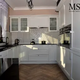 Jasna kuchnia z białymi szafkami z frezowanymi frontami, czarnym blatem i parkietem ułożonym w jodełkę; na blacie czajnik i garnek, w tle błyszczące płytki w romby.