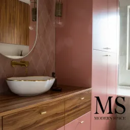 Nowoczesna łazienka z umywalką nablatową w białej misie ze złotym rantem, armaturą w kolorze starego złota, szafką z drewnianym blatem i różowymi frontami oraz wysoką szafą w tym samym kolorze.