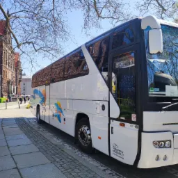 Biały autokar turystyczny z kolorowym logo na tle ceglanego budynku uniwersyteckiego i drzew bez liści.