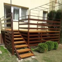 Brązowy taras z drewnianymi schodami, metalową balustradą i konstrukcją pergoli, przylegający do budynku z oknami, otoczony zielenią.