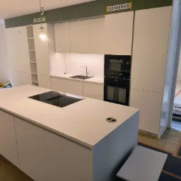 Wnętrze nowoczesnej kuchni z białą zabudową meblową, wyspą kuchenną z płytą indukcyjną i gniazdkami, widoczne elementy instalacji oświetleniowej i wentylacyjnej, podłoga częściowo zabezpieczona folią.