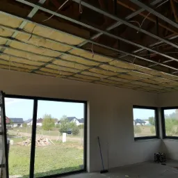 Poddasze w trakcie ocieplania wełną mineralną umieszczoną w metalowej konstrukcji, widoczne okna i drabina oparta o ścianę.