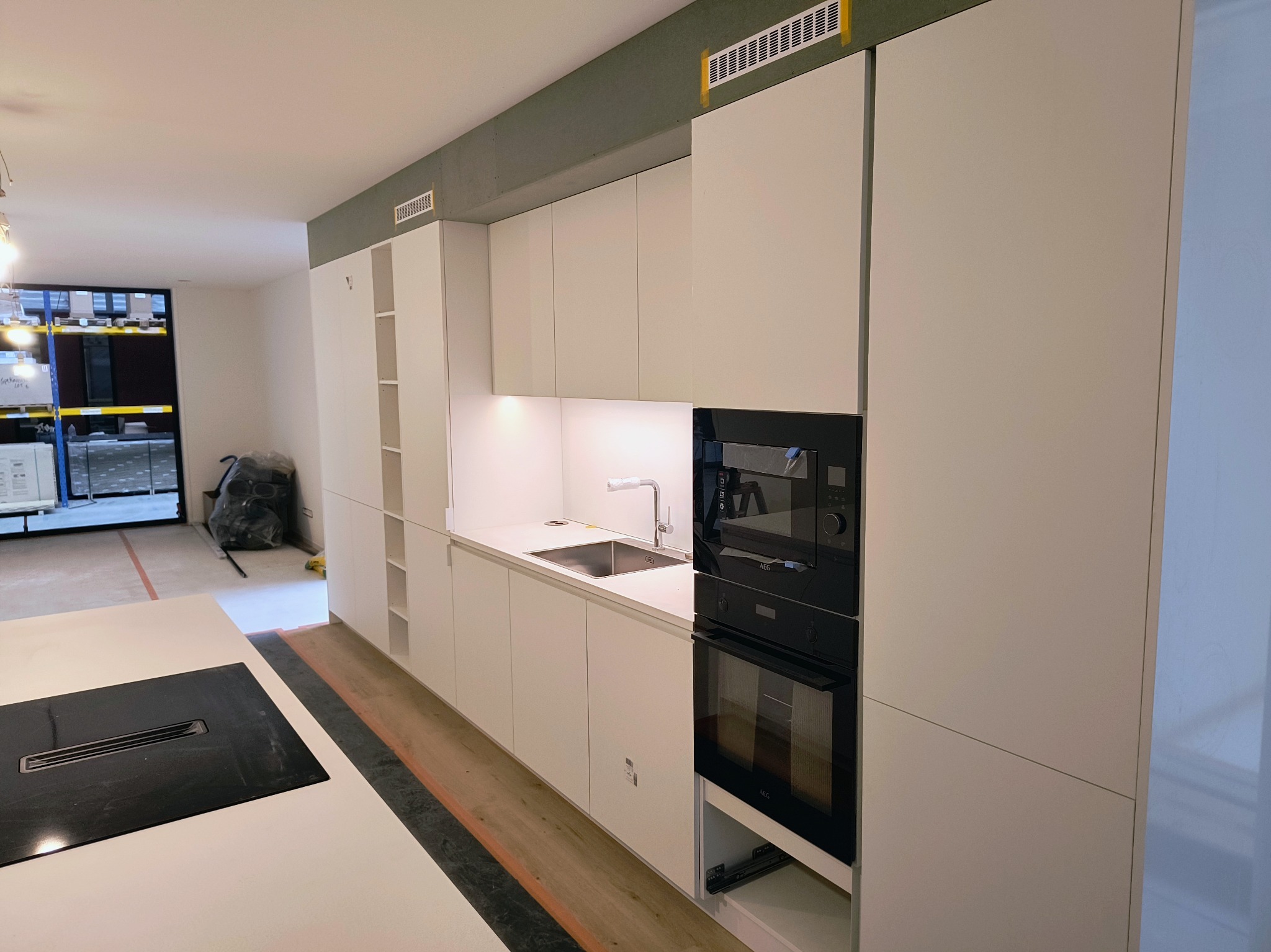 Nowoczesna, jasna kuchnia w trakcie montażu, widoczne białe szafki kuchenne, czarny piekarnik i płyta indukcyjna, zabezpieczenia taśmą malarską na suficie i podłodze.