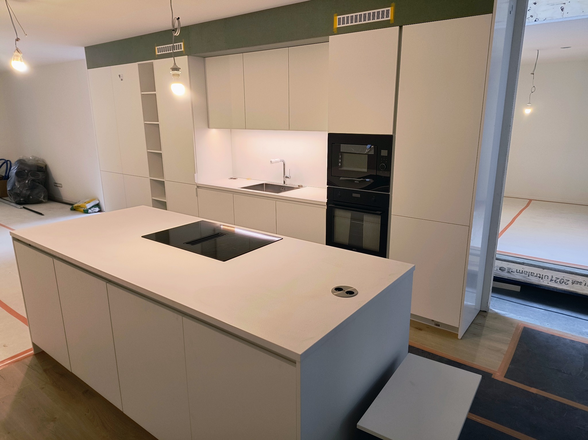 Wnętrze nowoczesnej kuchni z białą zabudową meblową, wyspą kuchenną z płytą indukcyjną i gniazdkami, widoczne elementy instalacji oświetleniowej i wentylacyjnej, podłoga częściowo zabezpieczona folią.