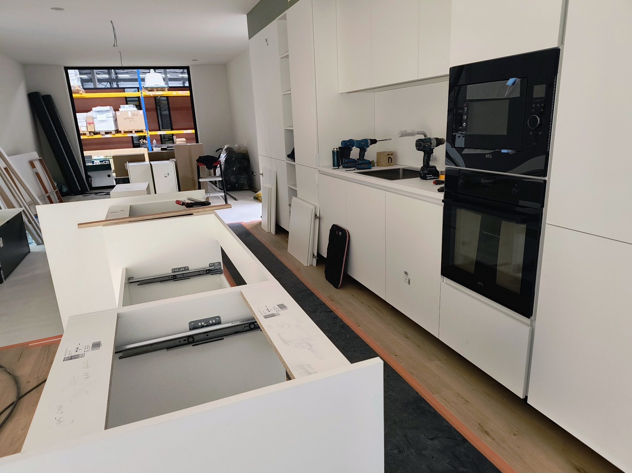 Montaż nowoczesnej białej kuchni z wyspą; widoczne szuflady z zamontowanymi prowadnicami, zabudowany piekarnik AEG, w tle narzędzia i okno na magazyn.