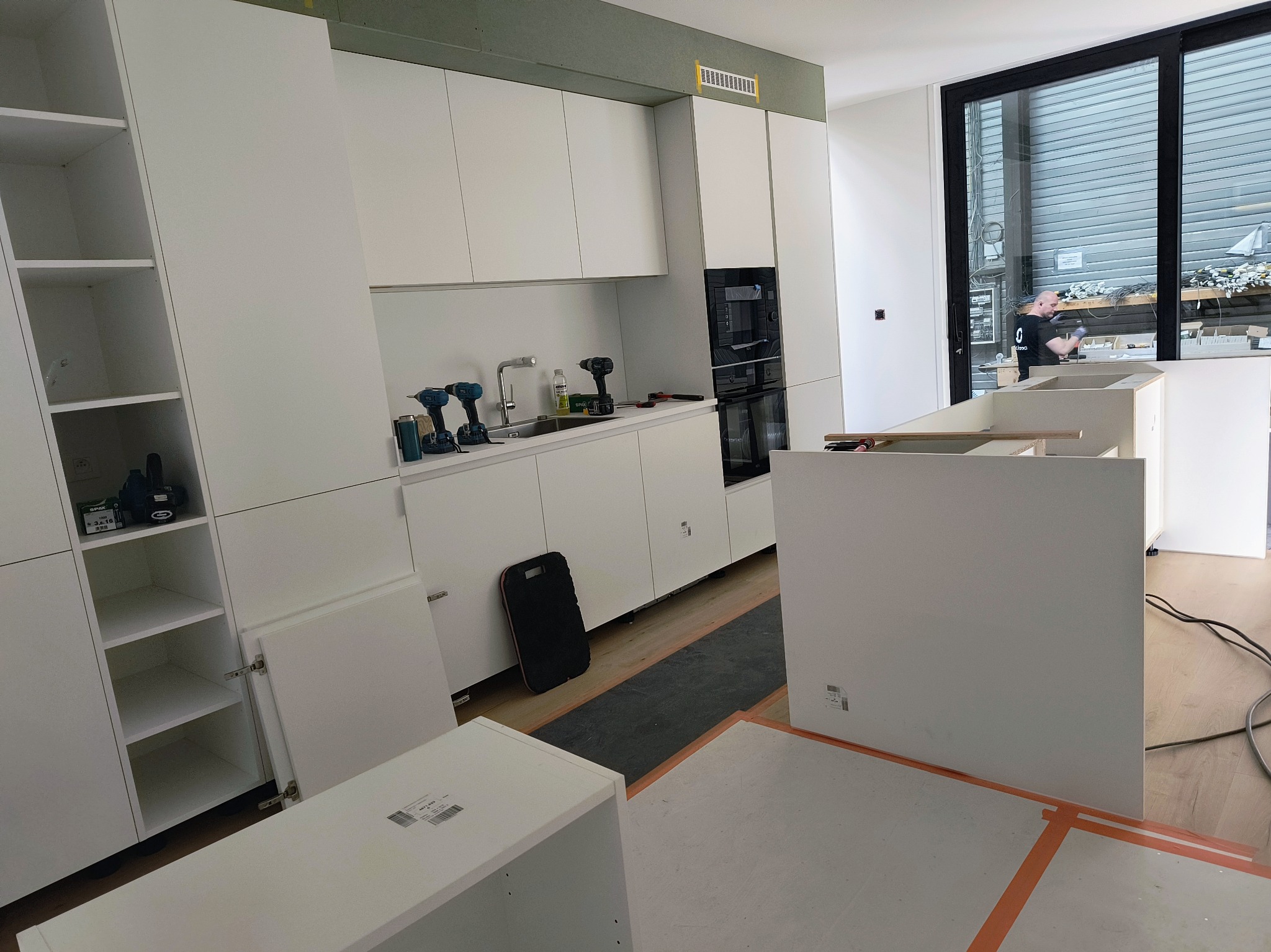 Montaż białej zabudowy kuchennej w surowym wnętrzu, widoczne narzędzia, części szafek i pracownik w tle za oknem.