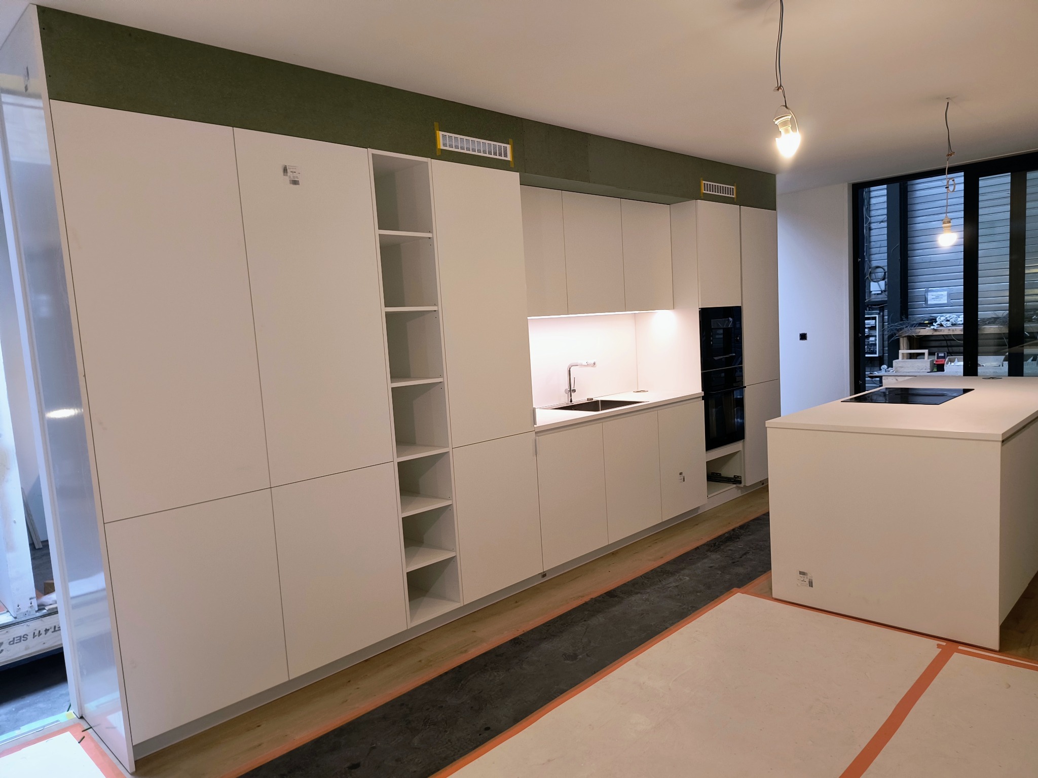 Nowoczesna biała kuchnia z wyspą, w trakcie prac wykończeniowych, widoczne zabezpieczone podłogi i surowe żarówki.