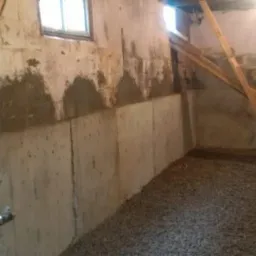 Piwnica z częściowo naprawionymi ścianami fundamentowymi z betonu, podpartymi drewnianymi belkami, z podsypką żwirową na podłodze i małym oknem u góry.