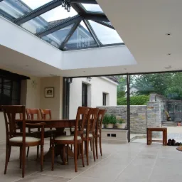 Jasne wnętrze domu z przeszklonym dachem i ścianami, widok na kamienny mur i ogród, stół z krzesłami w centrum, podłoga z jasnych płyt.