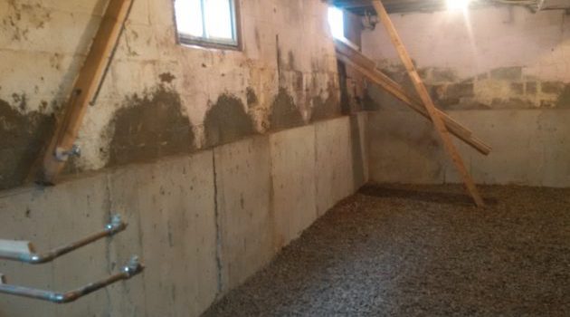 Piwnica z częściowo naprawionymi ścianami fundamentowymi z betonu, podpartymi drewnianymi belkami, z podsypką żwirową na podłodze i małym oknem u góry.