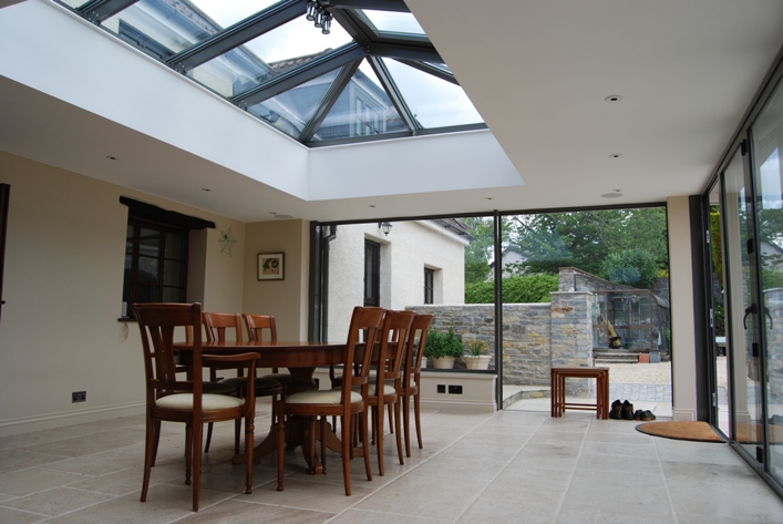 Jasne wnętrze domu z przeszklonym dachem i ścianami, widok na kamienny mur i ogród, stół z krzesłami w centrum, podłoga z jasnych płyt.