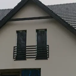Fragment elewacji domu z czarną, poziomą balustradą balkonową, okna zabezpieczone folią, dach pokryty ciemną dachówką