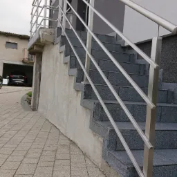 Zewnętrzne schody z szarej kostki brukowej prowadzące do budynku, zabezpieczone metalową, minimalistyczną barierką z rurek.