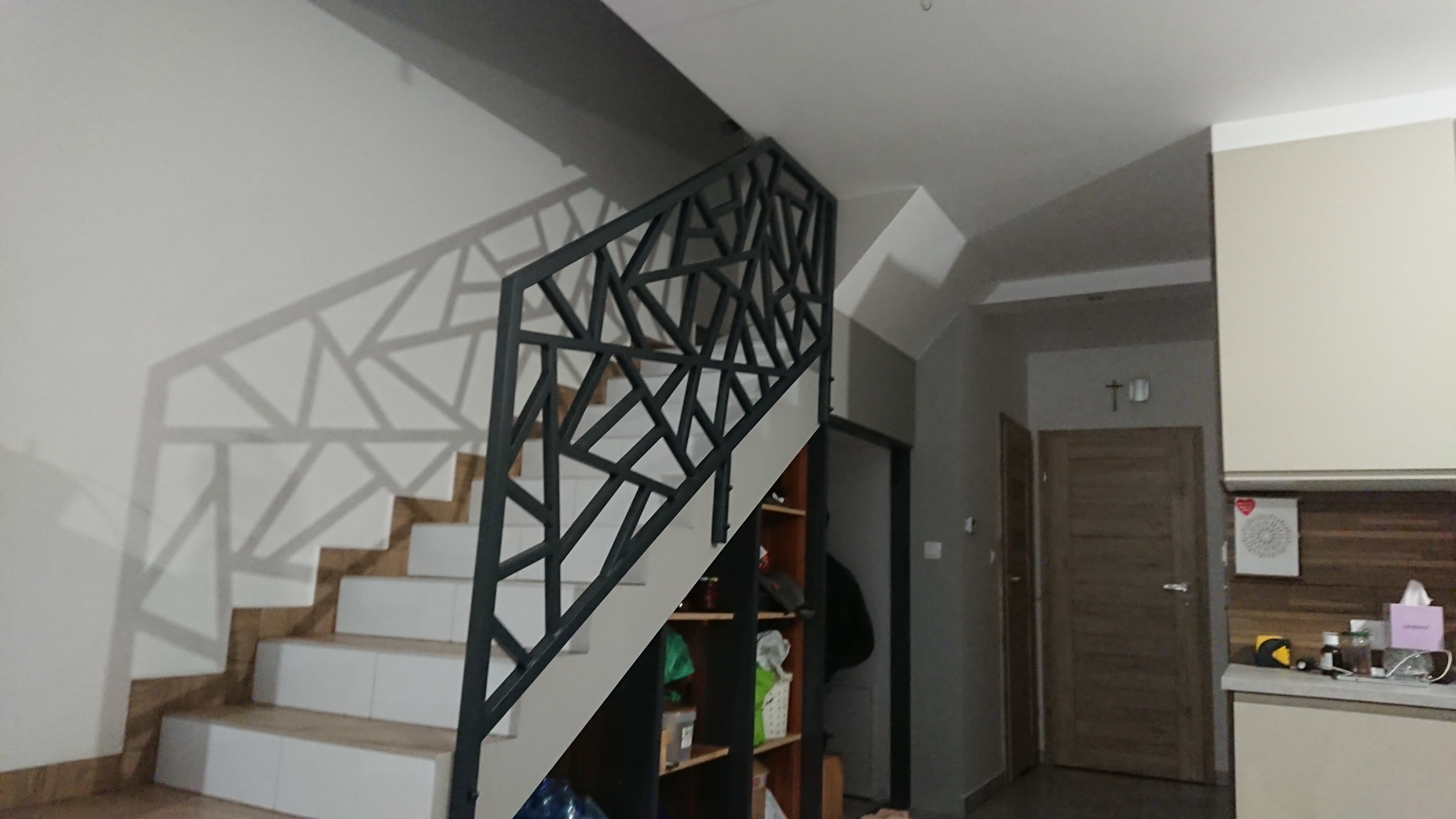 Nowoczesna, geometryczna balustrada schodowa w kolorze grafitowym, rzucająca cienie na białą ścianę, z widocznymi stopniami schodów i fragmentem regału w tle.