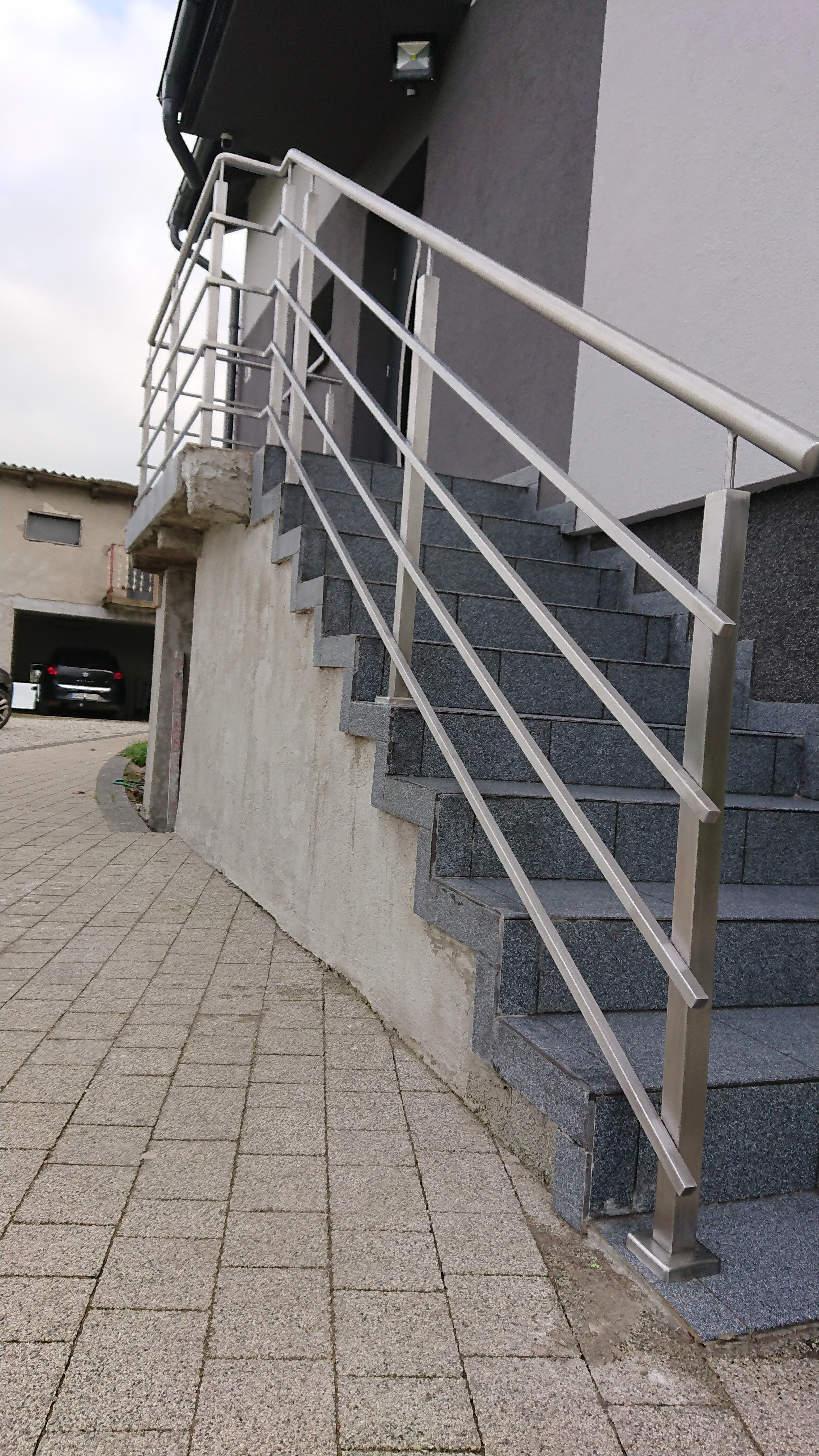 Zewnętrzne schody z szarej kostki brukowej prowadzące do budynku, zabezpieczone metalową, minimalistyczną barierką z rurek.