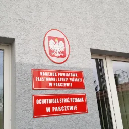 Tablice informacyjne na elewacji budynku Komendy Powiatowej Państwowej Straży Pożarnej i Ochotniczej Straży Pożarnej w Parczewie, z godłem Polski w okrągłej obwódce.