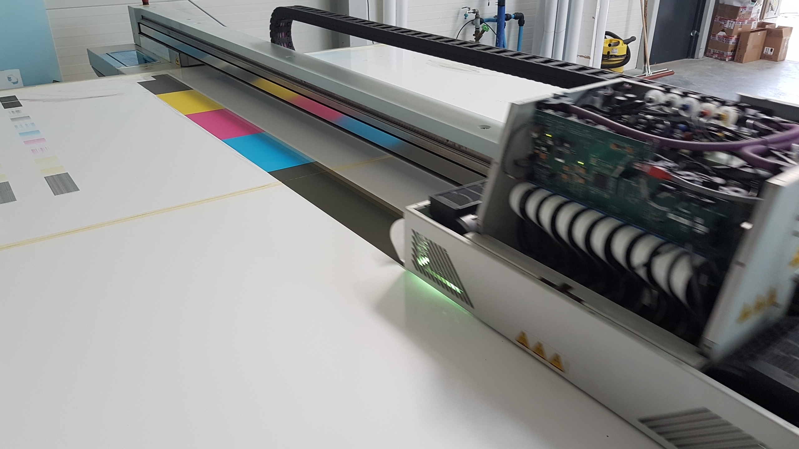 Drukarka wielkoformatowa w trakcie kalibracji kolorów, widoczne paski testowe CMYK na białym podłożu, z otwartym panelem bocznym ukazującym elektronikę i przewody.