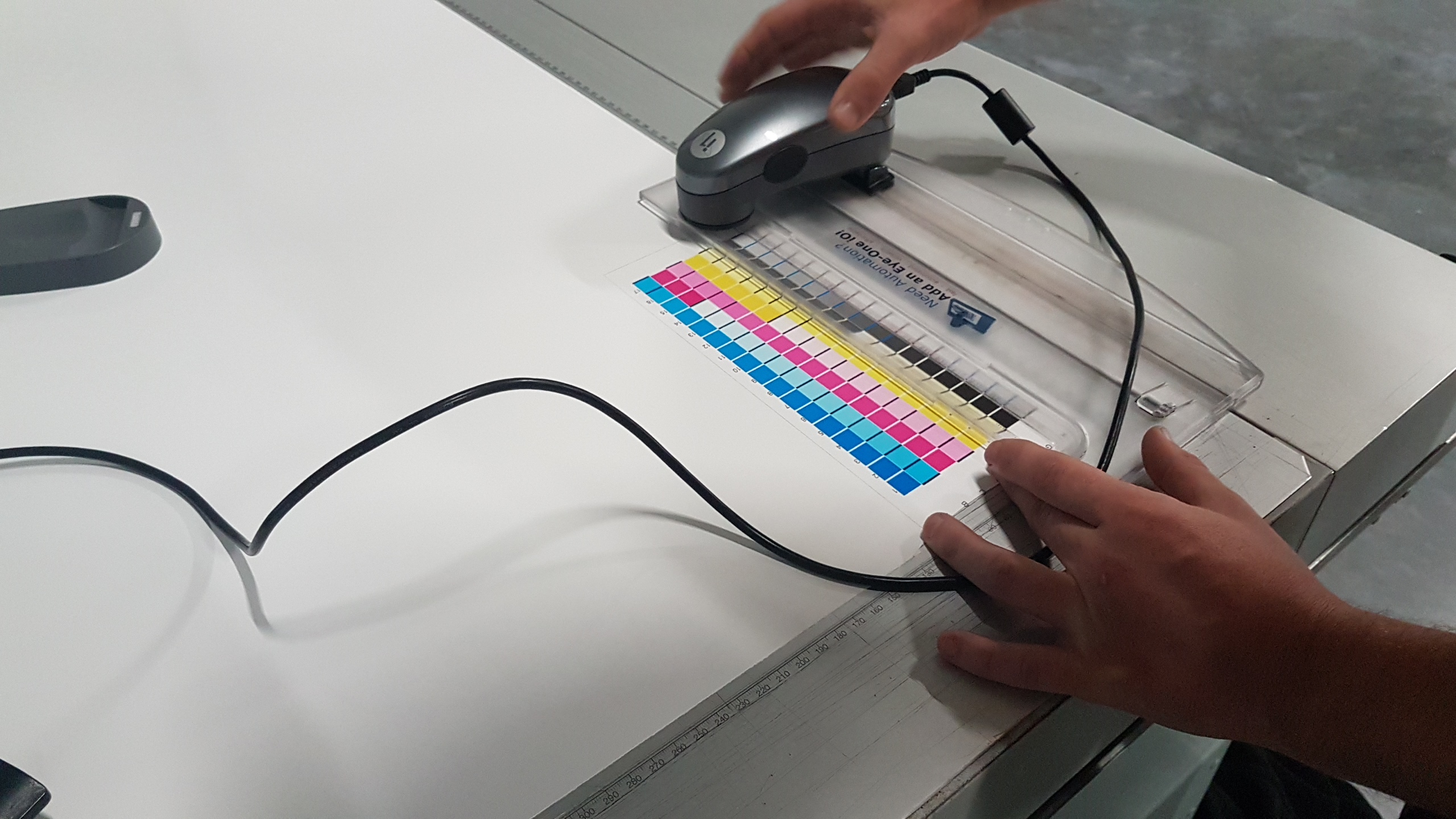 Kalibracja kolorów na wydruku za pomocą spektrofotometru Eye-One. Kolorowe pola testowe, linijka i ręce operatora na białym stole.