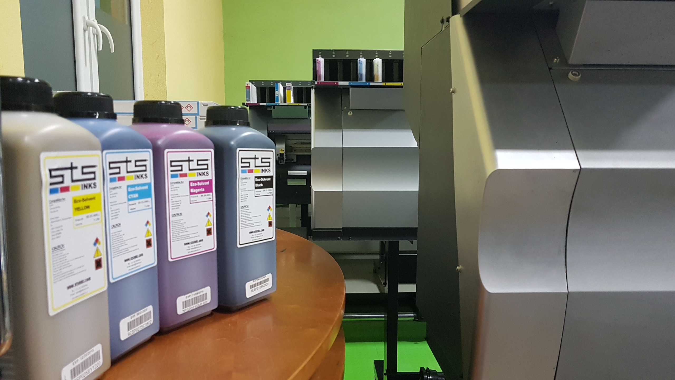 Butelki z tuszami Eco-Solvent w kolorach yellow, cyan, magenta i black marki STS INKS, ustawione na drewnianym stole, z widoczną drukarką wielkoformatową w tle.