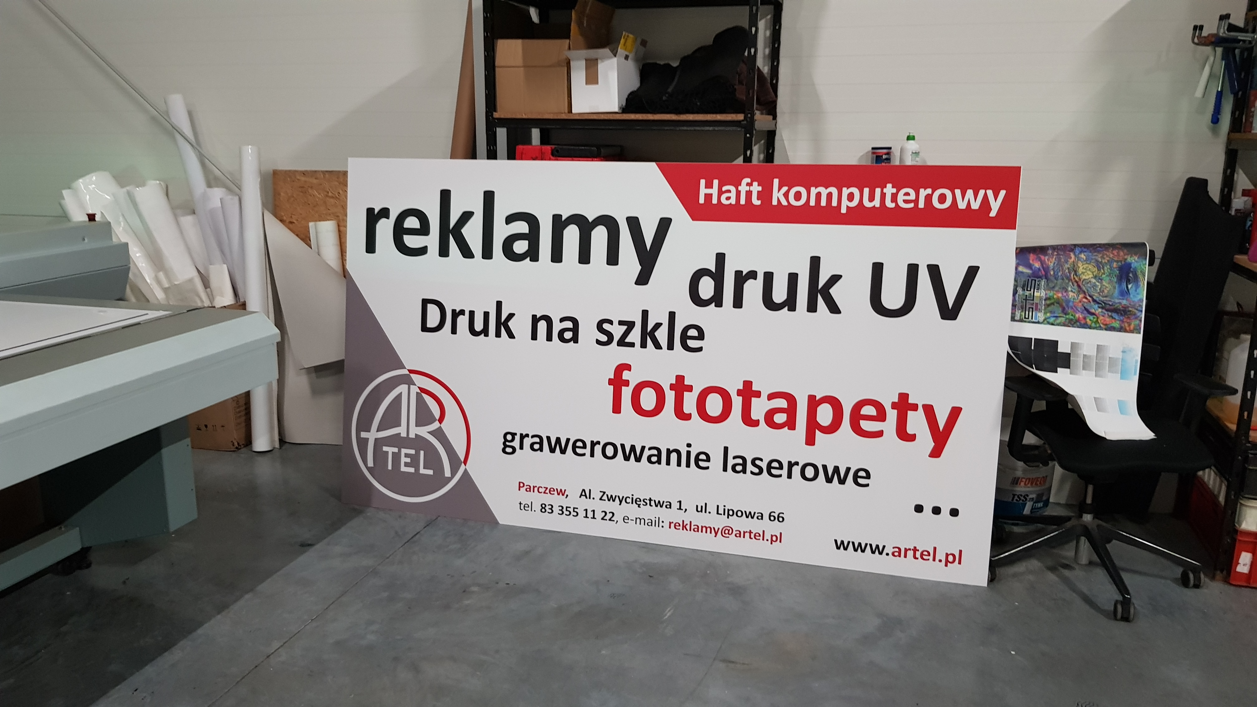 Duży baner reklamowy z napisem 'reklamy', 'druk UV', 'fototapety' i danymi kontaktowymi firmy Artel, ustawiony w warsztacie drukarskim, obok maszyny drukarskiej i regałów z materiałami.
