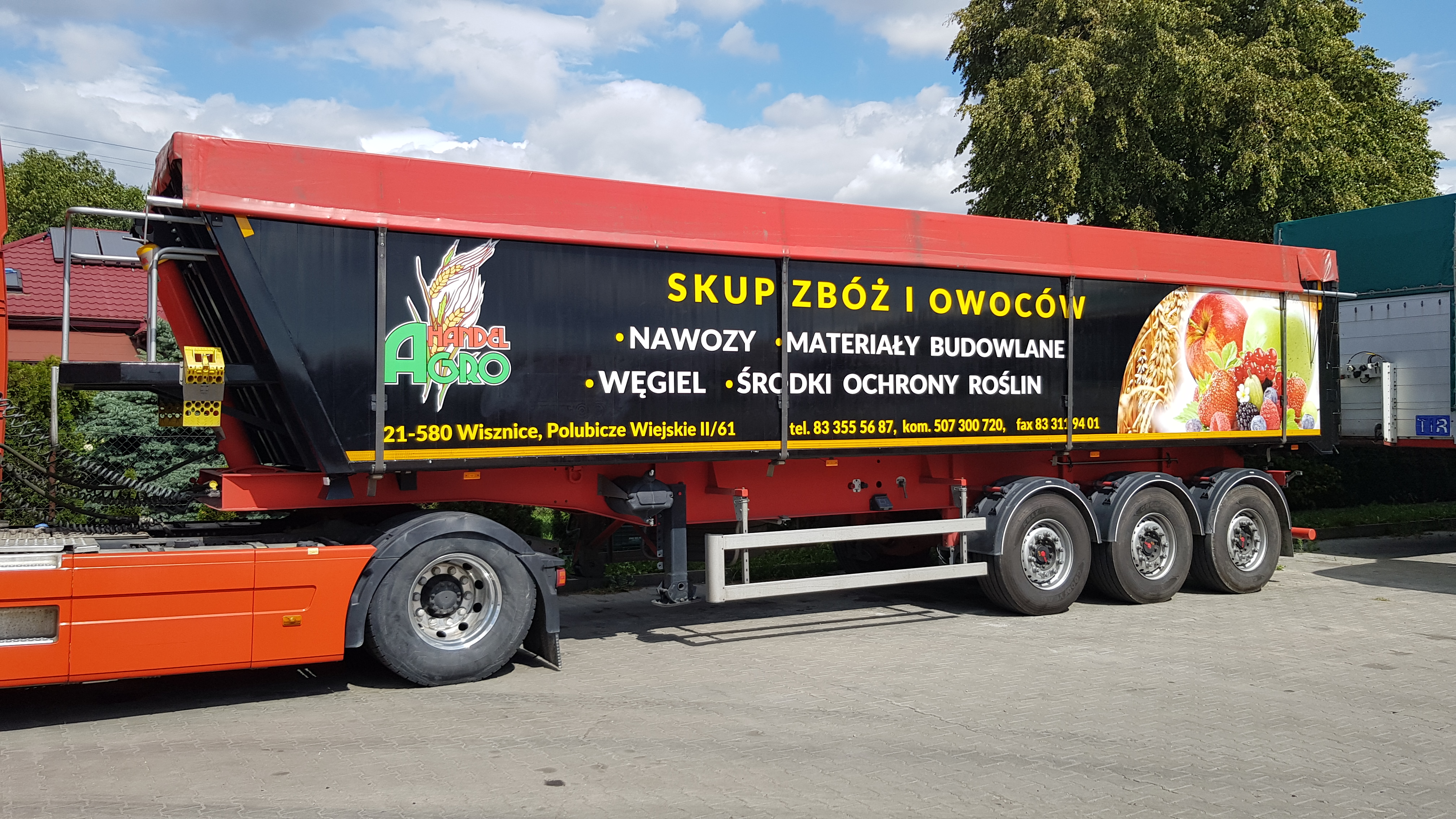Czerwono-czarna naczepa ciężarówki z reklamą skupu zbóż i owoców, nawozów, materiałów budowlanych, węgla i środków ochrony roślin, zaparkowana na placu.