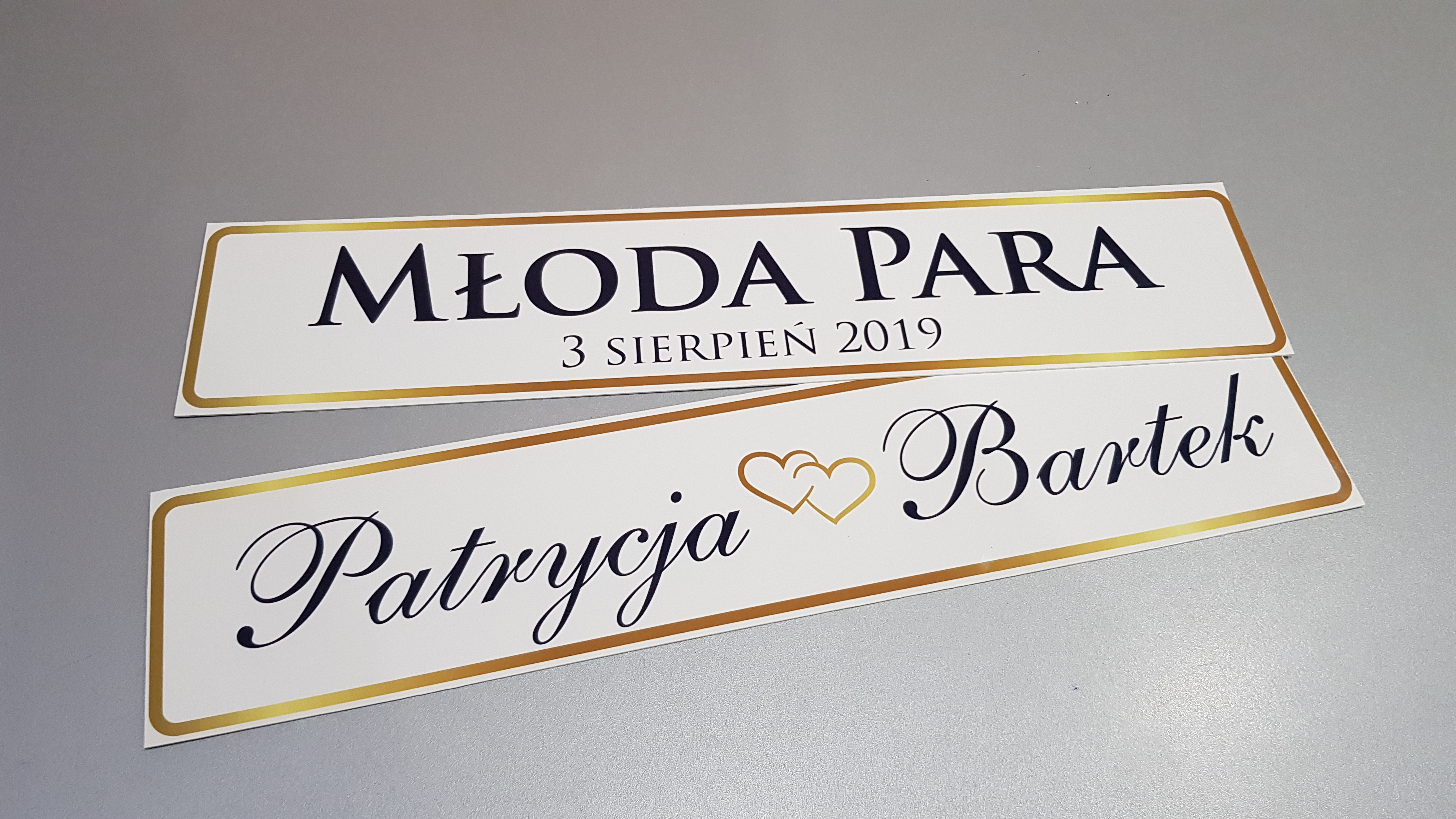 Dwa białe, prostokątne szyldy z czarnym napisem i złotą obwódką: górny z napisem 'Młoda Para, 3 Sierpień 2019', dolny z imionami 'Patrycja' i 'Bartek' rozdzielonymi dwoma złotymi sercami, leżące...