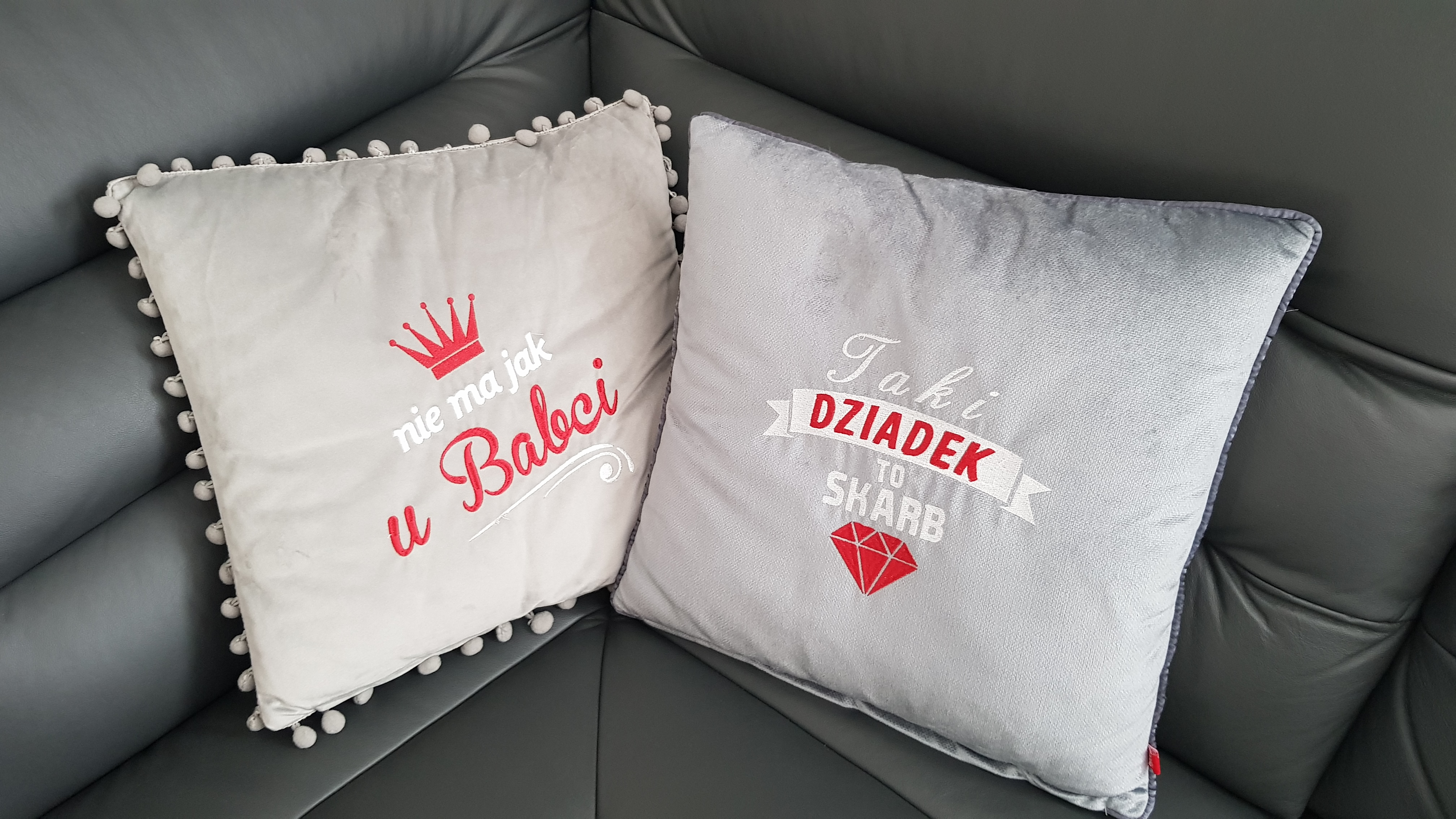 Dwie dekoracyjne poduszki z haftowanymi napisami 'nie ma jak u Babci' z koroną i 'Taki Dziadek to Skarb' z diamentem, leżące na ciemnoszarej skórzanej sofie.