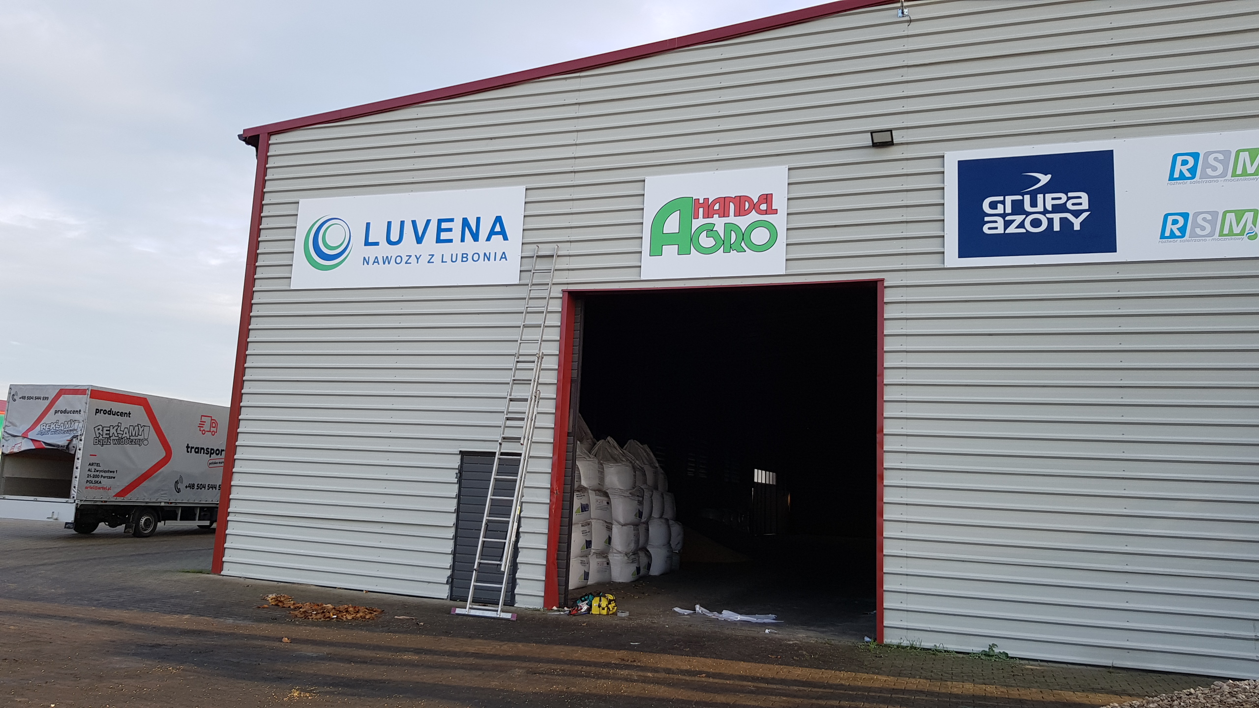 Fasada magazynu z zamontowanymi tablicami reklamowymi firm Luvena, Handel Agro i Grupa Azoty, drabina oparta o ścianę, w tle zaparkowana ciężarówka z reklamą.