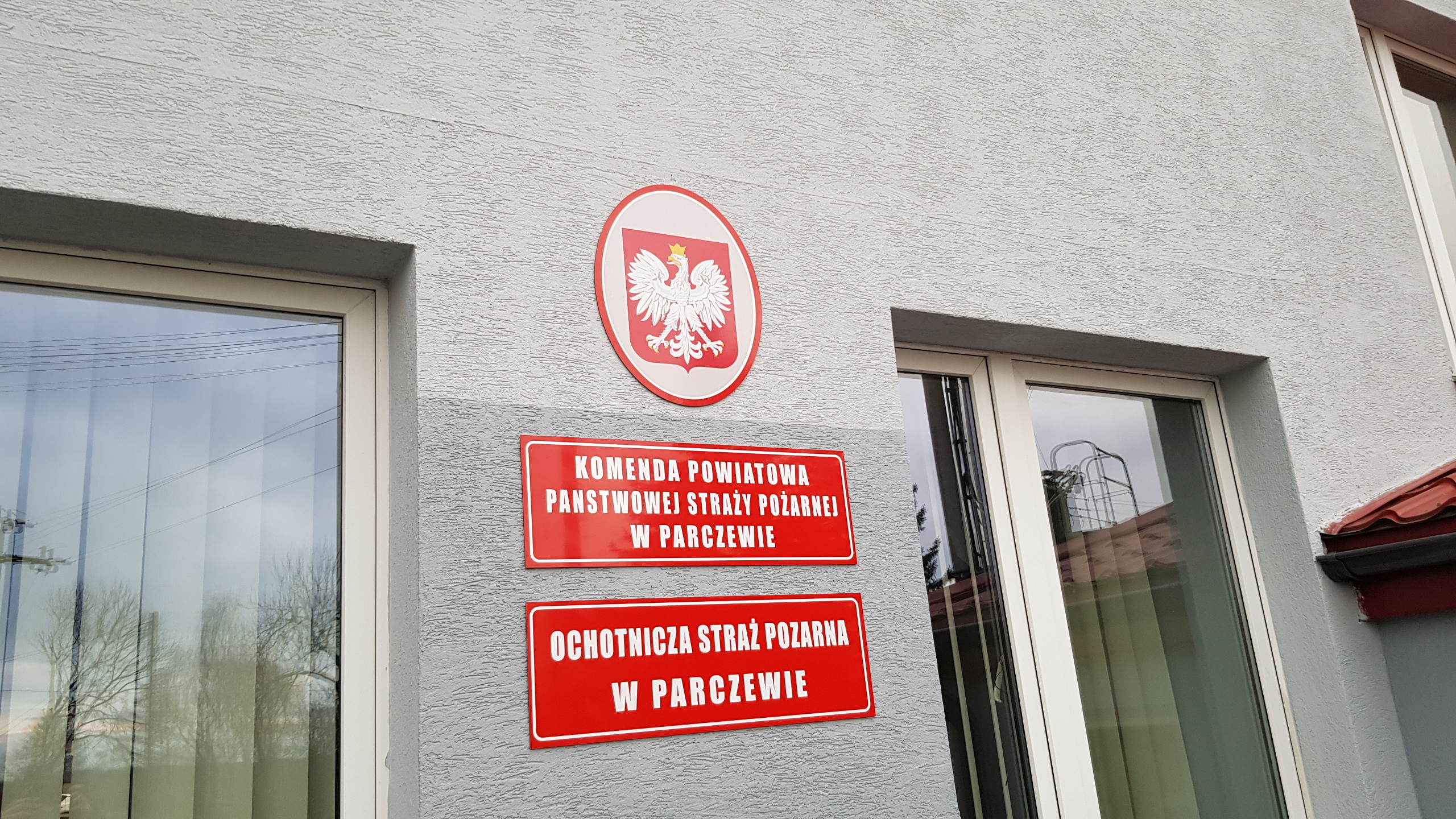 Tablice informacyjne na elewacji budynku Komendy Powiatowej Państwowej Straży Pożarnej i Ochotniczej Straży Pożarnej w Parczewie, z godłem Polski w okrągłej obwódce.