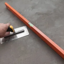 Ręka z pacą wygładza szary betonowy podkład, obok poziomnica murarska.