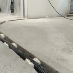 Czarna rura karbowana ułożona w wyżłobionym kanale w szarej betonowej podłodze, zabezpieczona pianką montażową. W tle widoczna szlifierka kątowa Makita i częściowo zasłonięte folią ściany.