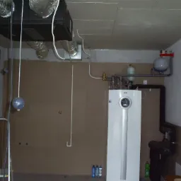 Biały, nowoczesny bojler elektryczny z podłączeniami hydraulicznymi w kotłowni, widoczne rury i instalacja, w tle surowy, niezakończony sufit podwieszany i aluminiowe przewody wentylacyjne.