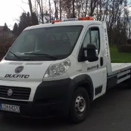 Biały Fiat Ducato z platformą do transportu pojazdów, zaparkowany na asfalcie z widocznymi pomarańczowymi światłami ostrzegawczymi na dachu i numerem rejestracyjnym LCH 64370.