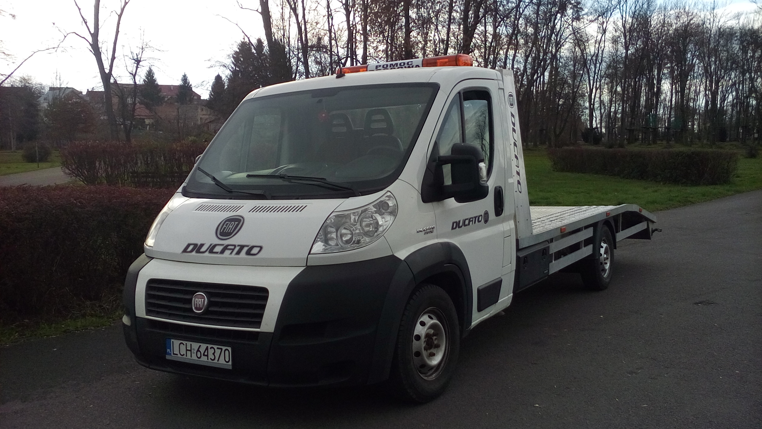 Biały Fiat Ducato z platformą do transportu pojazdów, zaparkowany na asfalcie z widocznymi pomarańczowymi światłami ostrzegawczymi na dachu i numerem rejestracyjnym LCH 64370.