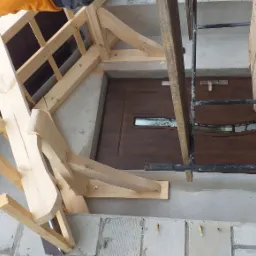 Surowa konstrukcja drewniana nad wejściem, widoczne elementy więźby dachowej, fragment elewacji z drabiną i brązowe drzwi wejściowe w tle.