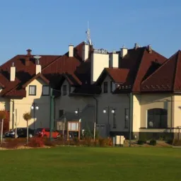 Duży budynek z brązowym dachem wielospadowym, kremową elewacją i licznymi kominami, widoczny z trawnika w słoneczny dzień.