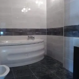 Nowoczesna łazienka z biało-szarymi płytkami, ciemnym pasem dekoracyjnym, owalną wanną, umywalką z czarną szafką i ciemną podłogą z płytek imitujących marmur.