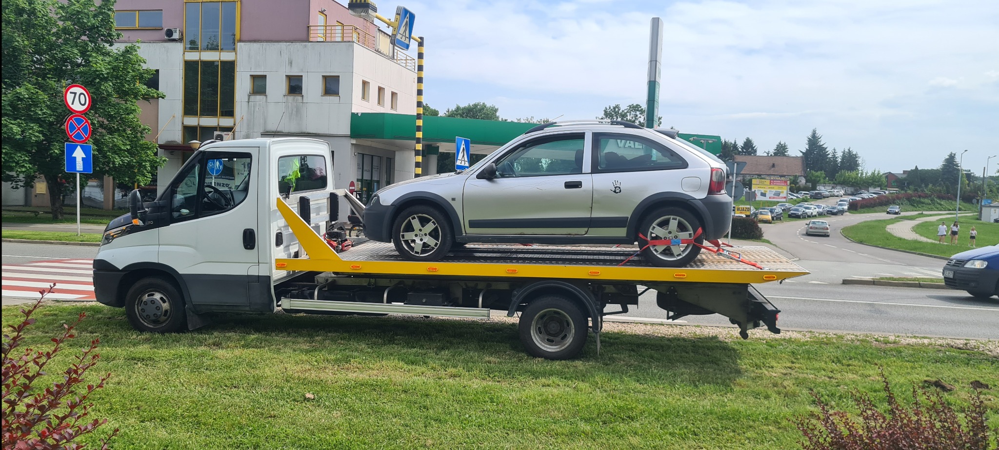 Laweta z srebrnym autem osobowym na żółtej platformie, zabezpieczonym czerwonymi pasami. W tle budynek z zielonymi elementami i znak drogowy.