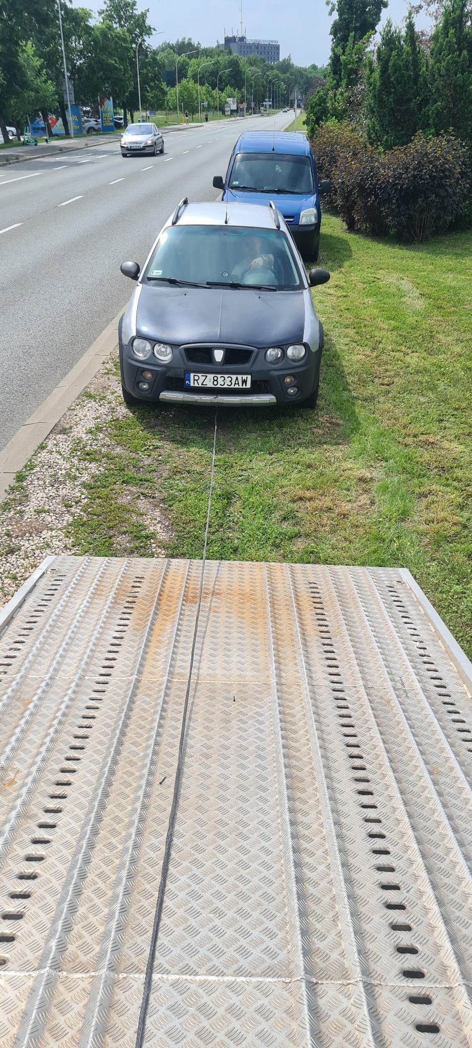 Szary Rover z numerem RZ 833AW holowany na lawecie. W tle zaparkowany niebieski van i ruchliwa ulica w słoneczny dzień. Widok z tyłu lawety.