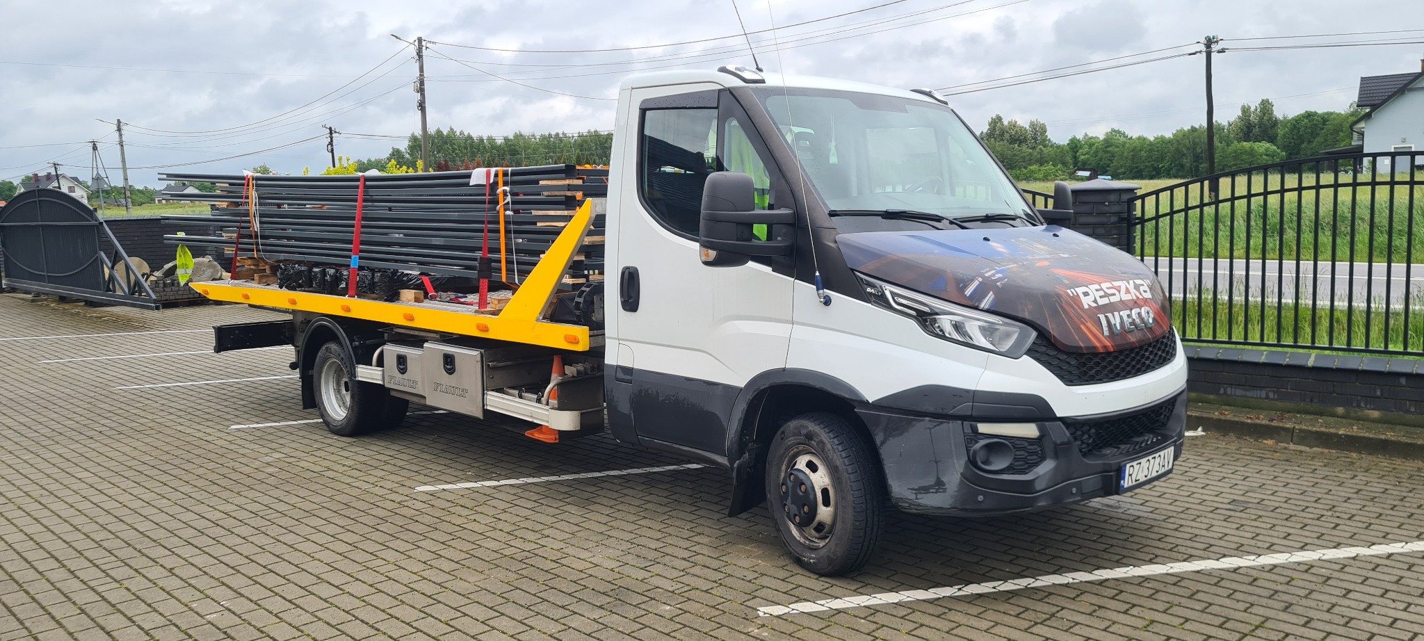 Laweta z ładunkiem metalowych elementów ogrodzeniowych na parkingu, z widoczną bramą w tle. Na masce pojazdu grafika z napisem 'RESZKA IVECO'.