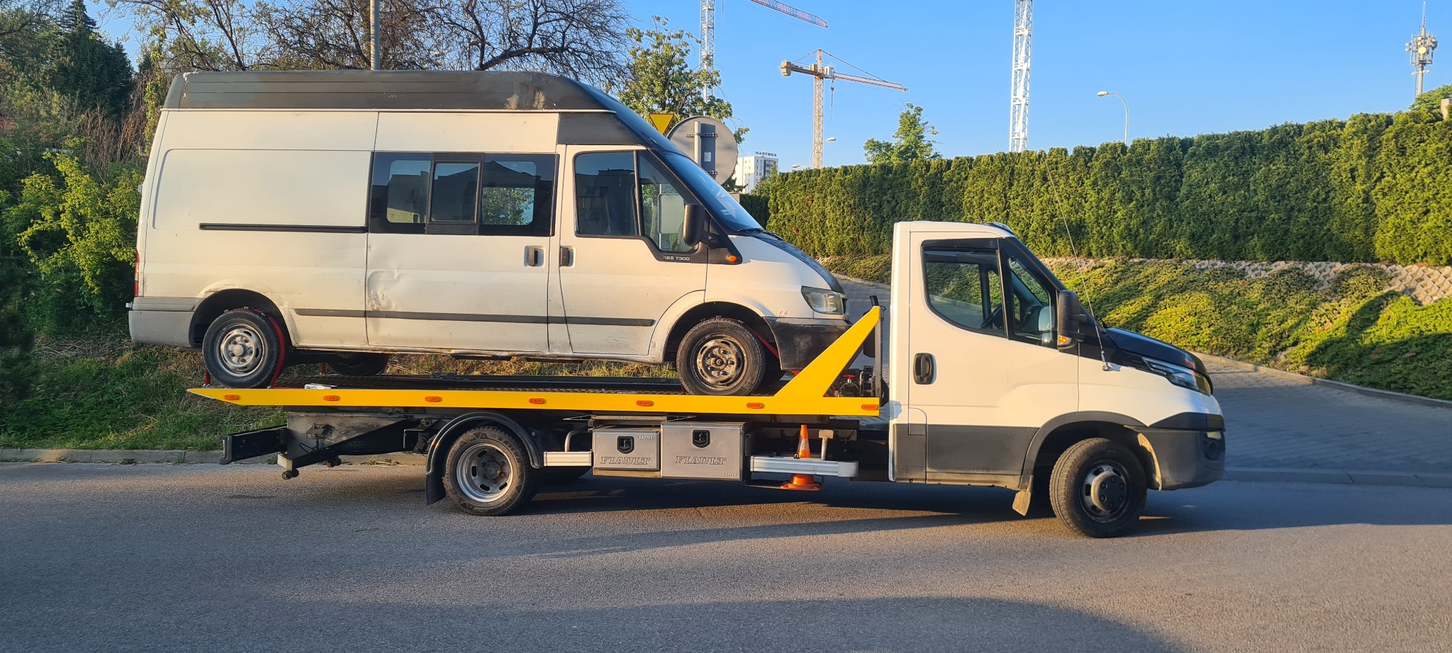Biała laweta z żółtym najazdem transportuje uszkodzony biały bus na tle zieleni i budynków. Widoczne detale: skrzynki narzędziowe i pachołki ostrzegawcze.