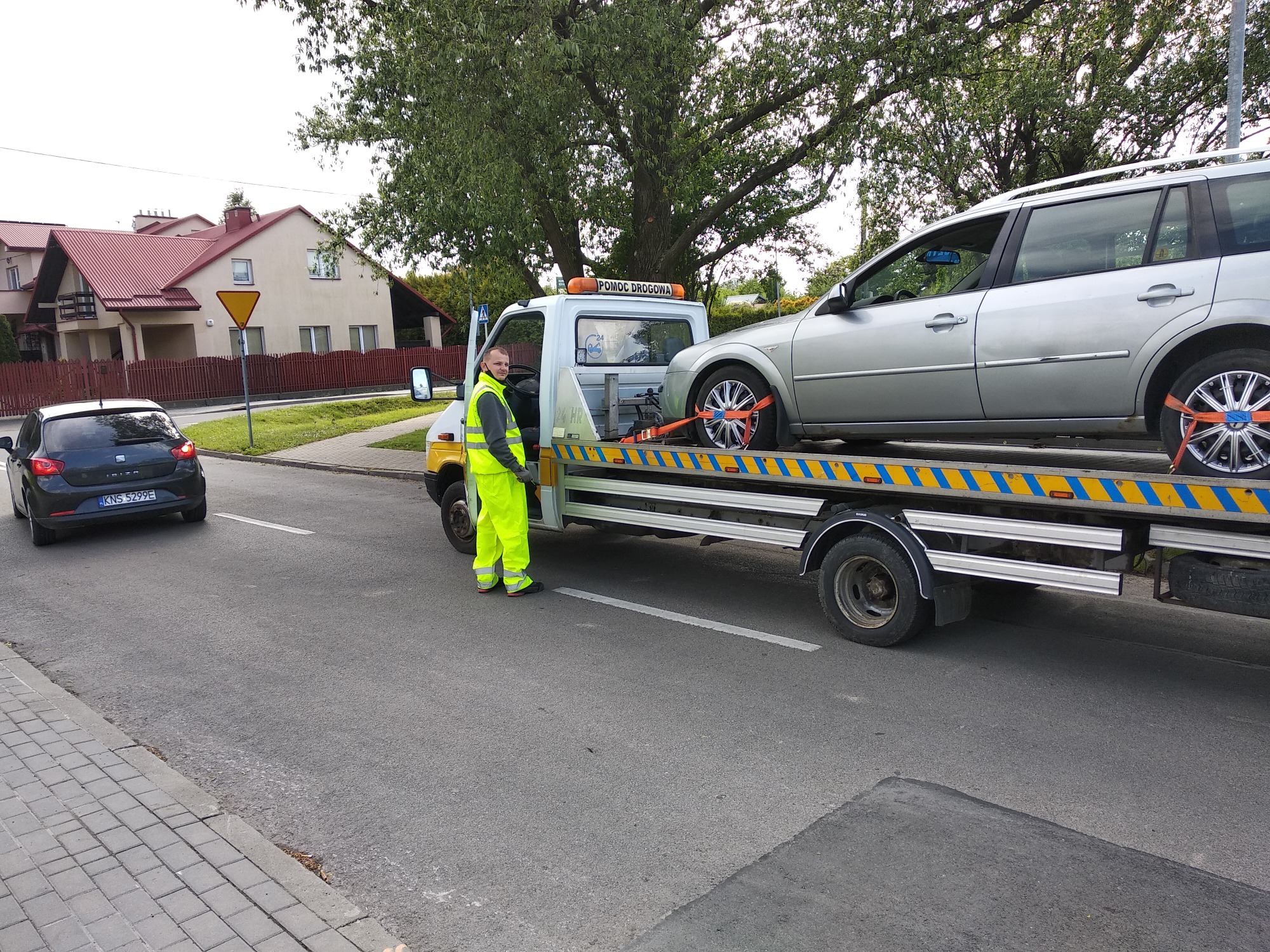 Laweta z załadowanym srebrnym kombi, zabezpieczonym pomarańczowymi pasami, pracownik w żółtym ubraniu stoi obok, w tle domy i drzewa.