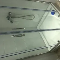 Szklana kabina prysznicowa z chromowanym uchwytem i baterią, widok z góry, z widocznym brodzikiem i fragmentem wykończenia ściany w płytkach imitujących drewno.