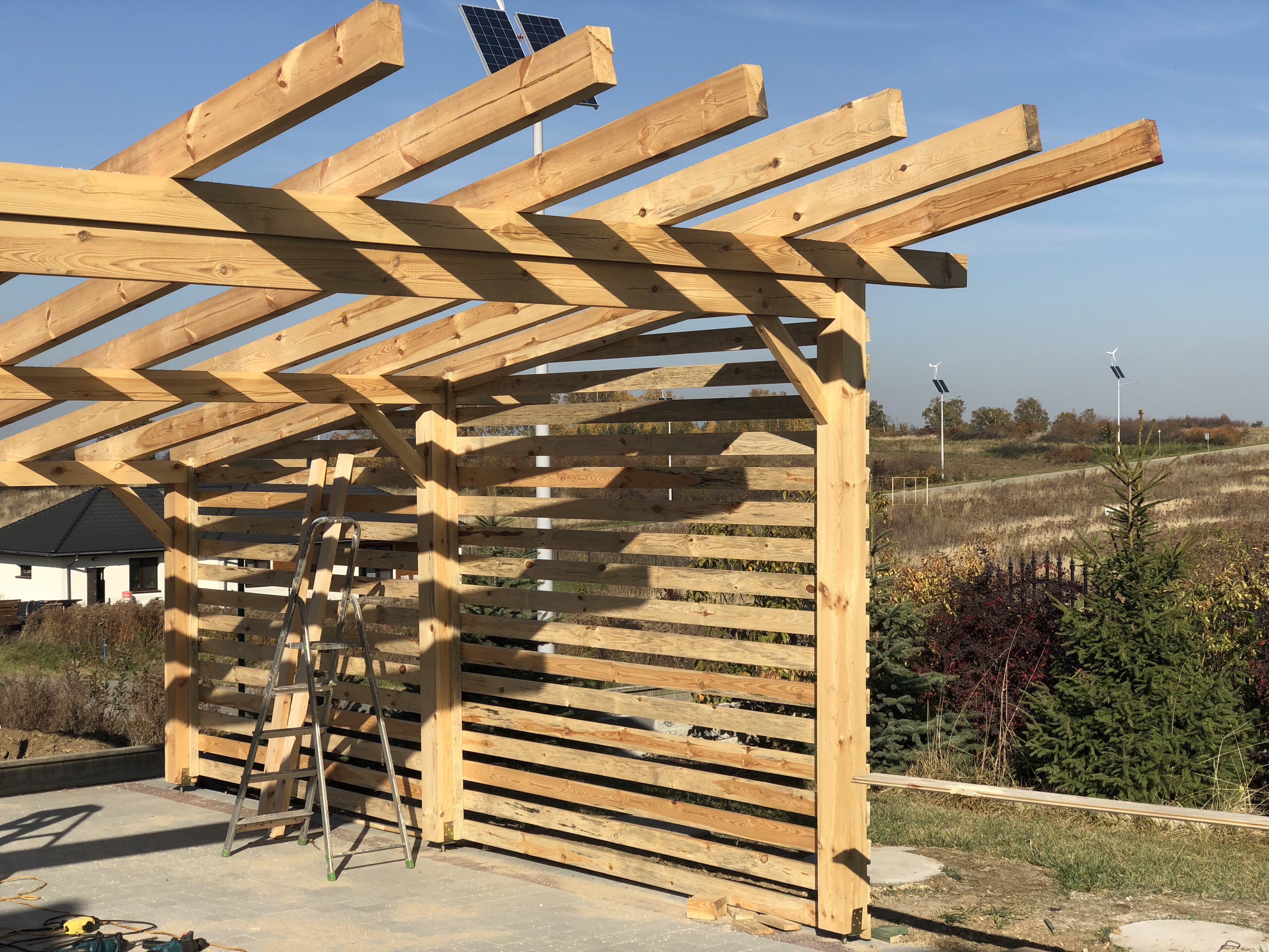 Drewniana konstrukcja altany w trakcie budowy, widoczna drabina i narzędzia, w tle dom, panele słoneczne i turbiny wiatrowe.