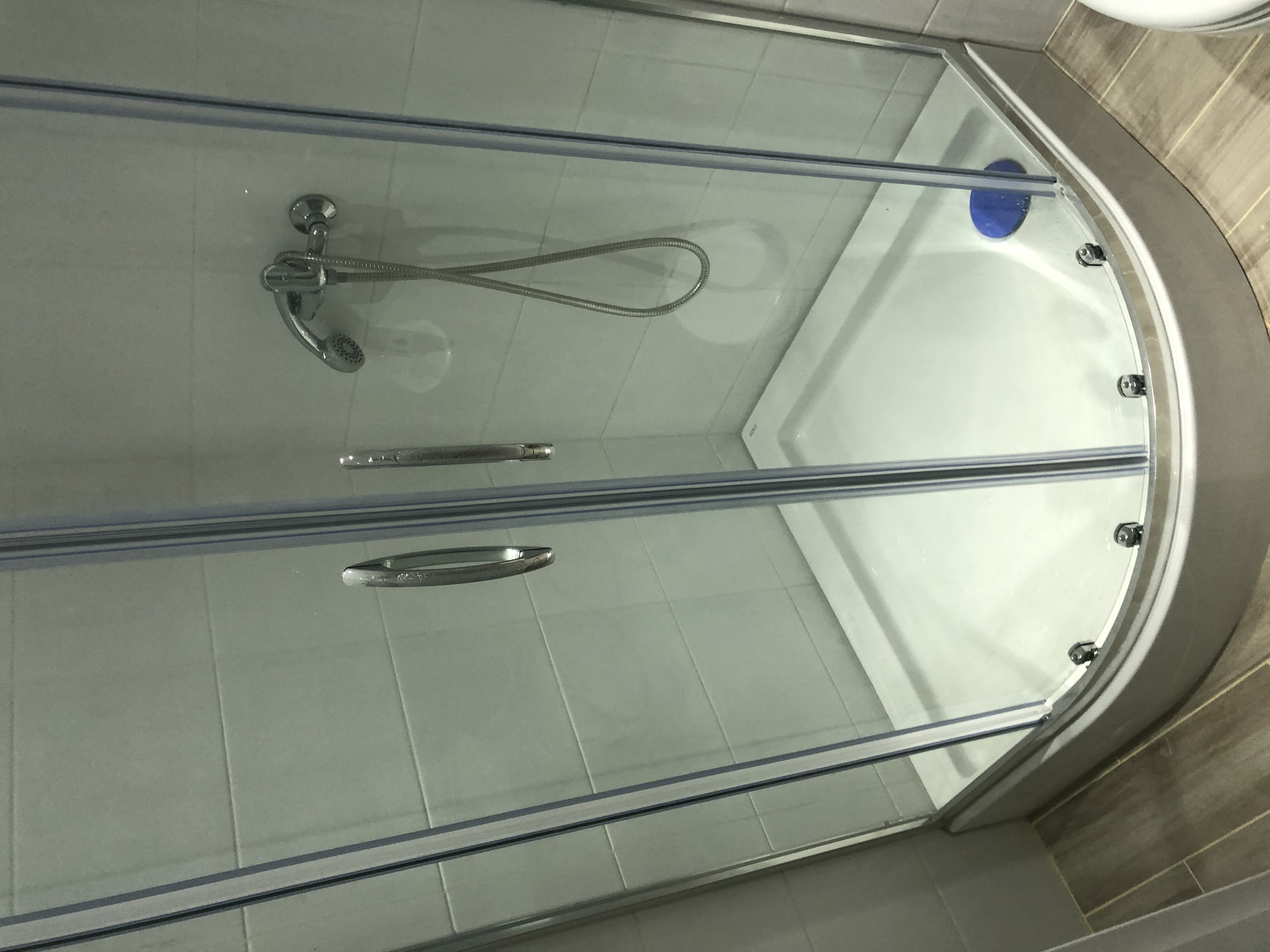 Szklana kabina prysznicowa z chromowanym uchwytem i baterią, widok z góry, z widocznym brodzikiem i fragmentem wykończenia ściany w płytkach imitujących drewno.