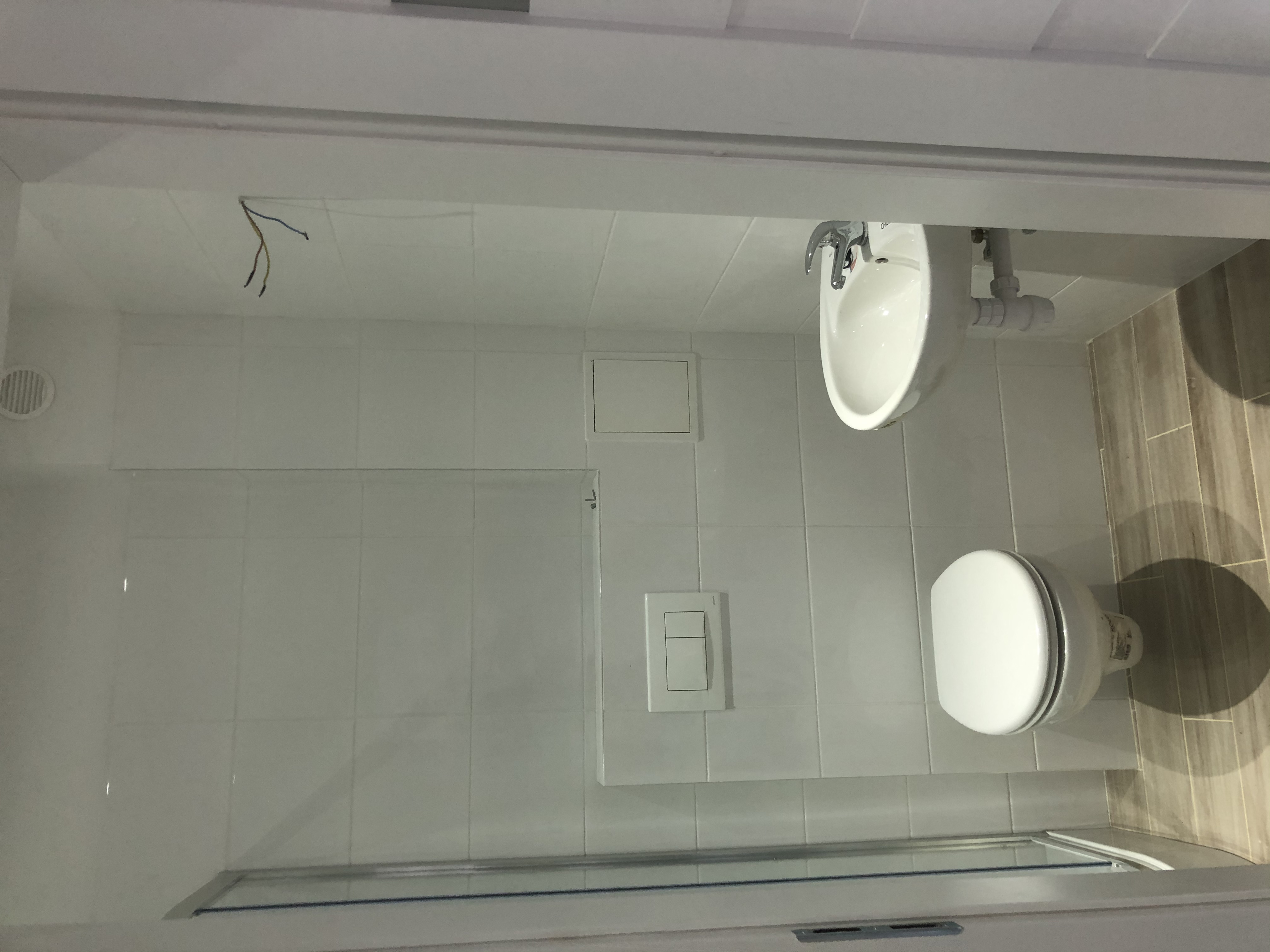 Biała, nowoczesna łazienka z umywalką, toaletą i płytkami imitującymi drewno; widoczne wystające przewody elektryczne.