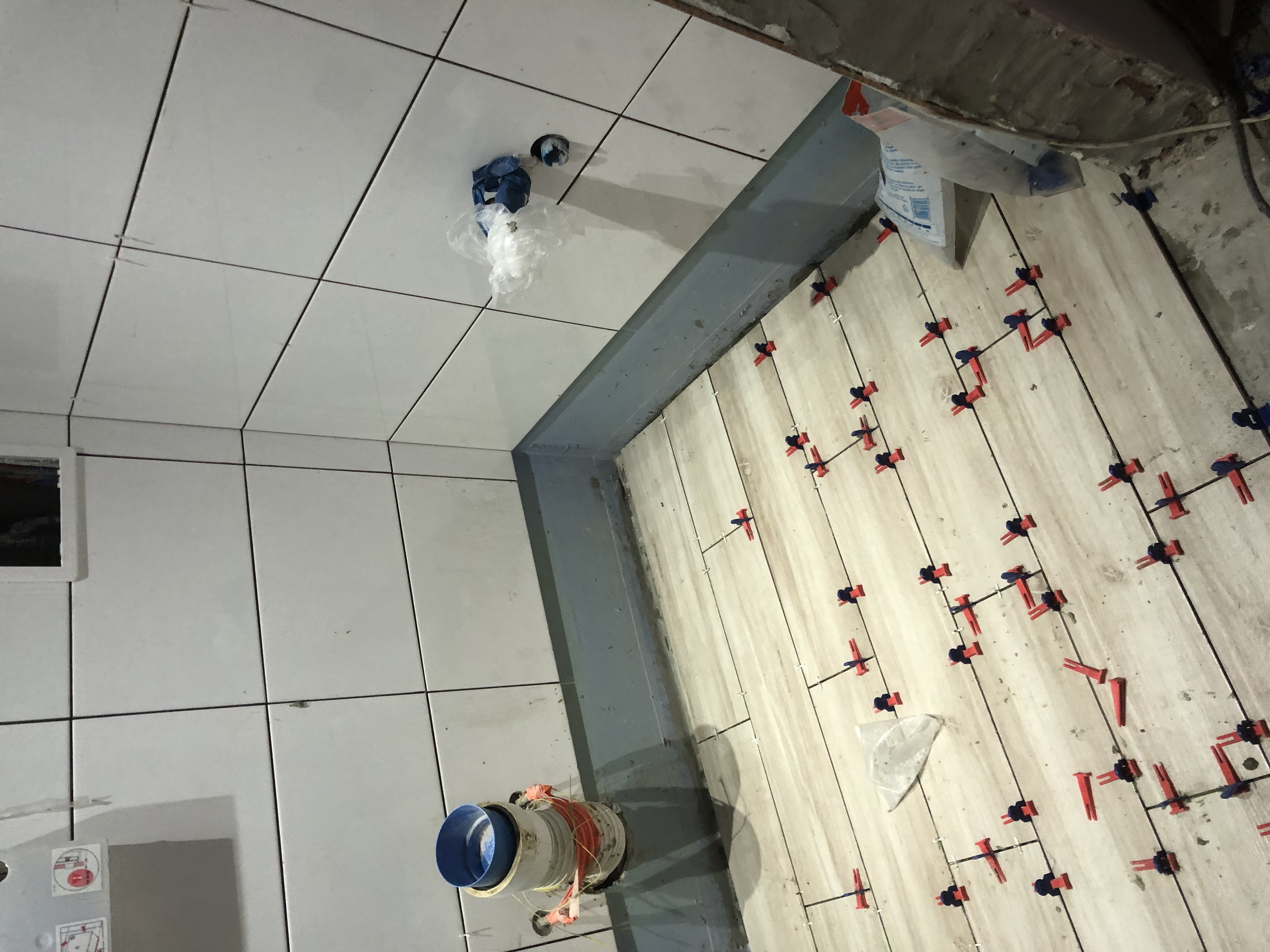 Układanie płytek podłogowych z użyciem systemu poziomowania: czerwone klipsy i niebieskie kliny, widoczne także rury kanalizacyjne i fragment ściany z płytkami.