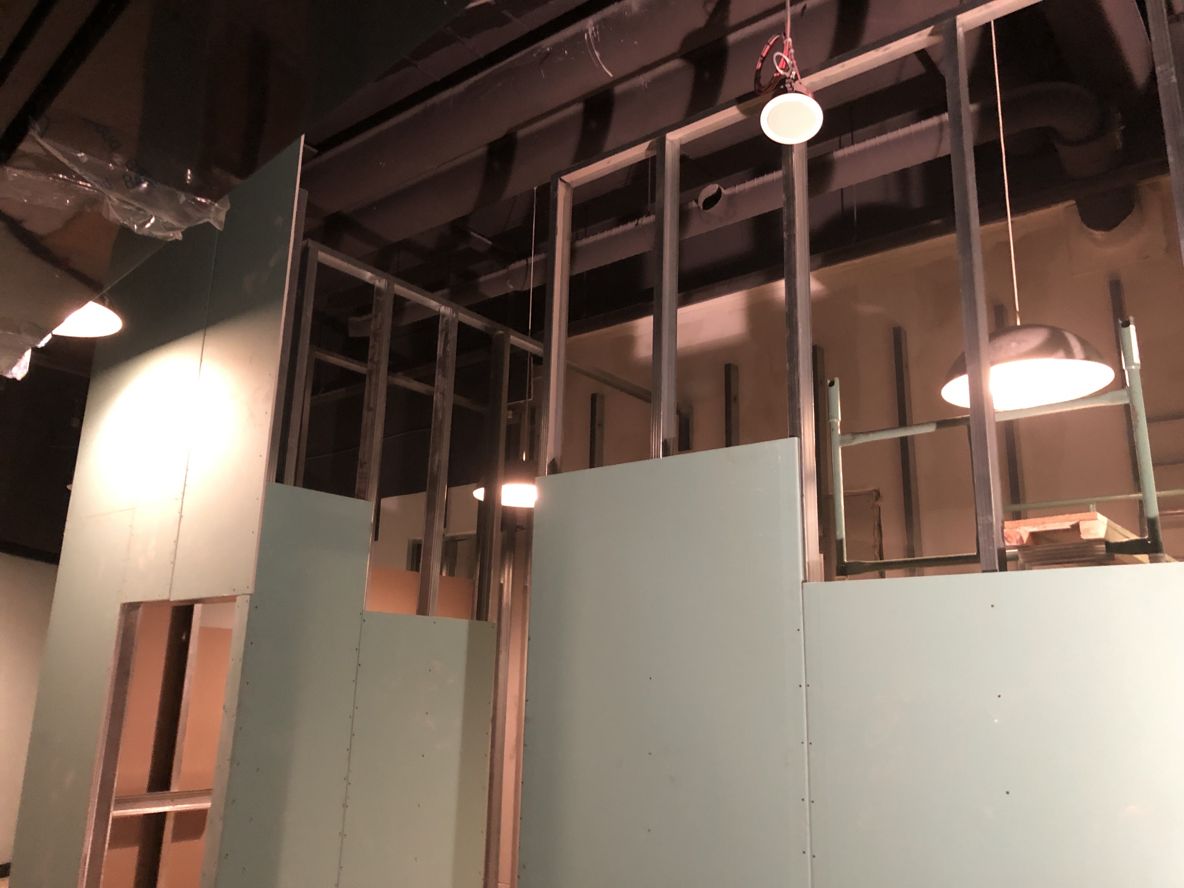 Wnętrze w trakcie prac: montaż płyt gipsowo-kartonowych na metalowym stelażu, widoczne instalacje pod sufitem i oświetlenie budowlane.